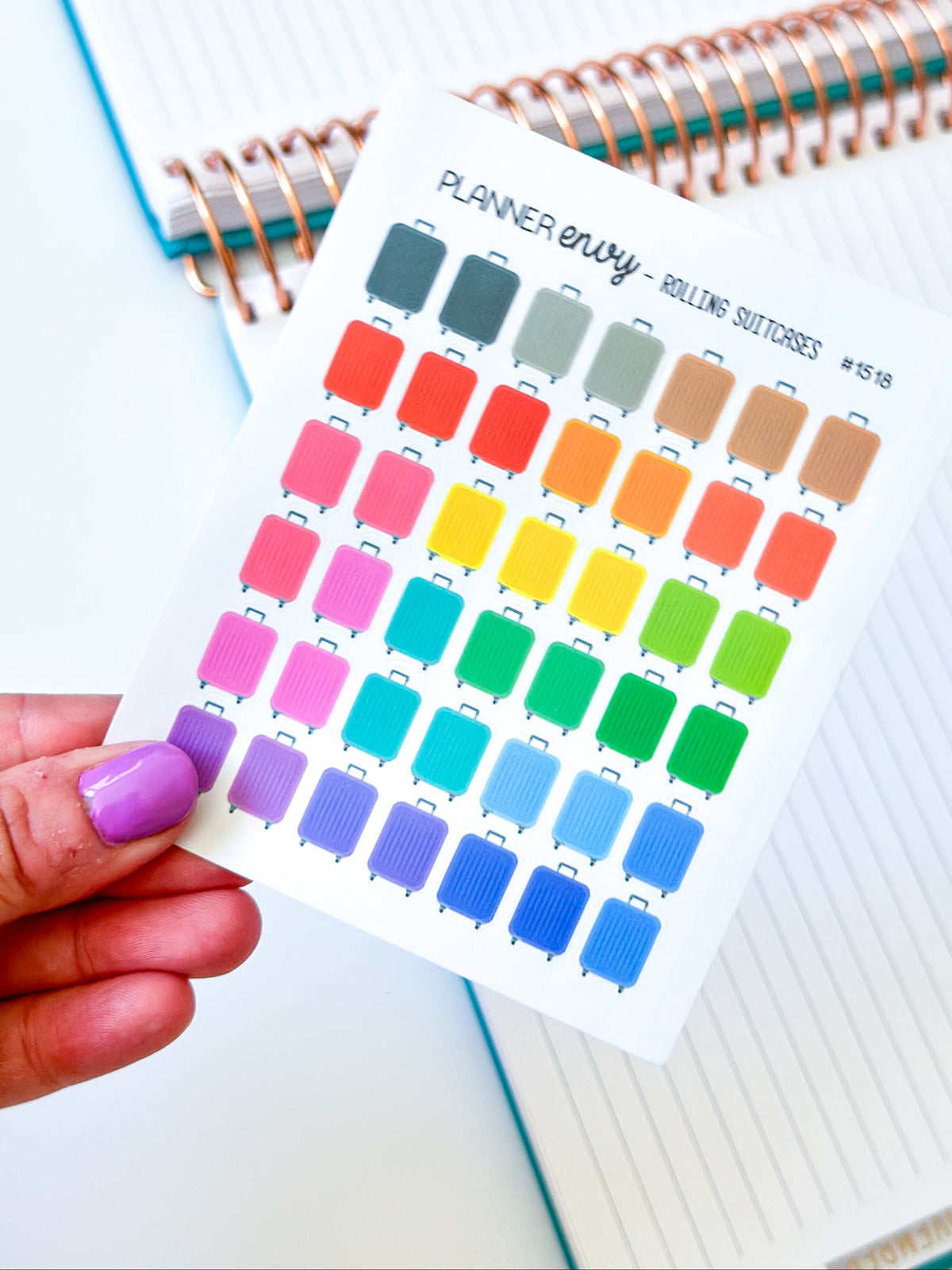 A hand with purple nail polish holds a sticker sheet featuring colorful luggage icons and labels for rolling suitcases, with lined notebooks in the background.